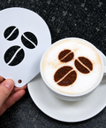 A hand holds a white metal stencil with three coffee bean cutouts next to a cup of cappuccino on a saucer, with the coffee topped to match the stencil shapes, on a dark speckled countertop.