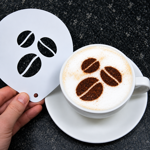 A hand holds a white metal stencil with three coffee bean cutouts next to a cup of cappuccino on a saucer, with the coffee topped to match the stencil shapes, on a dark speckled countertop.