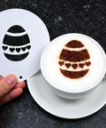 A hand holds a white circular stencil with a black Easter egg design next to a white cup of cappuccino on a saucer, showing the egg pattern created in the foam.