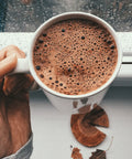 Hand holding a white mug filled with hot chocolate, with a blurred window background.