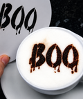 A white ceramic coffee cup filled with a white latte or cappuccino, topped with dark brown cocoa powder spelling 'BOO' in a dripping font. A hand is holding a matching white sticker or card with the same 'BOO' design against a dark textured background.