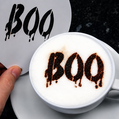 A white ceramic coffee cup filled with a white latte or cappuccino, topped with dark brown cocoa powder spelling 'BOO' in a dripping font. A hand is holding a matching white sticker or card with the same 'BOO' design against a dark textured background.