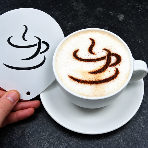 A hand holds a white ceramic coffee cup coaster with a black coffee cup graphic next to a white cup of cappuccino on a saucer, set against a dark textured background.