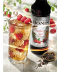 Glass of raspberry tea with ice and raspberries, Monin raspberry tea syrup bottle, and dried tea leaves on a white surface.
