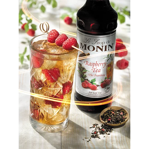 Glass of raspberry tea with ice and raspberries, Monin raspberry tea syrup bottle, and dried tea leaves on a white surface.