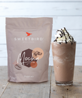 Sweetbird Mocha powder mix package next to a glass of mocha frappé with whipped cream on a wooden surface.