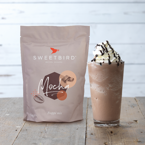 Sweetbird Mocha powder mix package next to a glass of mocha frappé with whipped cream on a wooden surface.