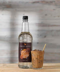 Bottle of Sweetbird Cane Syrup with a glass of iced coffee on a wooden surface.