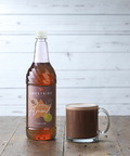 Bottle of Sweetbird Almond syrup next to a glass of hot chocolate on a wooden surface.