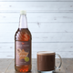 Bottle of Sweetbird Almond syrup next to a glass of hot chocolate on a wooden surface.