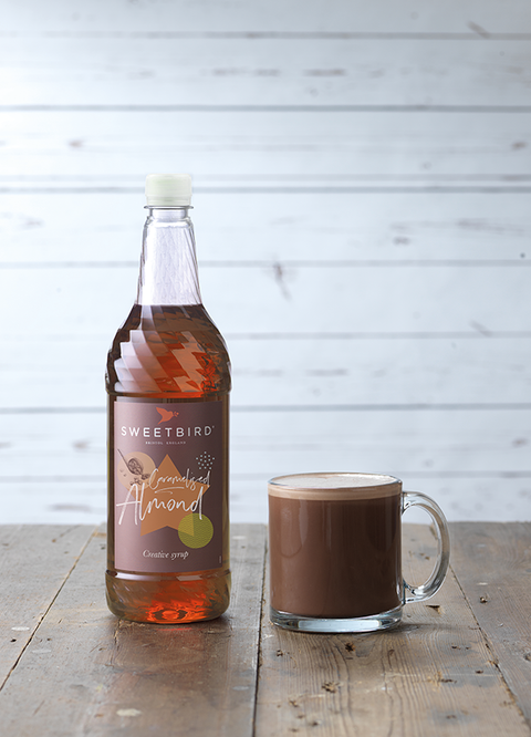 Bottle of Sweetbird Almond syrup next to a glass of hot chocolate on a wooden surface.