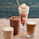 Three coffee drinks with whipped cream and a straw on a textured surface, featuring the Sweetbird brand.