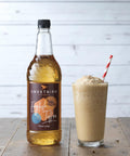 Bottle of Sweetbird English Toffee syrup next to a glass of a creamy drink on a wooden surface.
