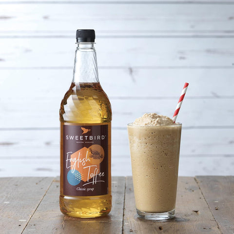 Bottle of Sweetbird English Toffee syrup next to a glass of a creamy drink on a wooden surface.