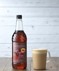 Bottle of Sweetbird Mac Pie syrup next to a cup of coffee on a wooden surface with a light gray background.
