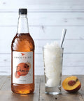 Bottle of Sweetbird Peach syrup with a glass of ice and a peach on a wooden surface.