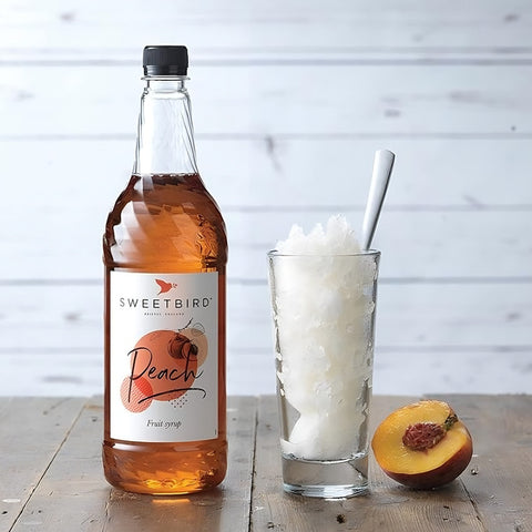Bottle of Sweetbird Peach syrup with a glass of ice and a peach on a wooden surface.