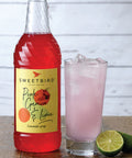 Bottle of Sweetbird Pink Guava & Lime lemonade syrup with a glass of pink lemonade and lime on a wooden surface.