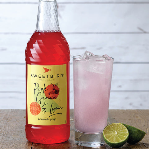 Bottle of Sweetbird Pink Guava & Lime lemonade syrup with a glass of pink lemonade and lime on a wooden surface.