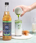 Person pouring a green liquid into a glass with a bottle of Tate & Lyle Barista Collection Hazelnut syrup.