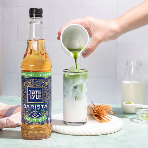 Person pouring a green liquid into a glass with a bottle of Tate & Lyle Barista Collection Hazelnut syrup.