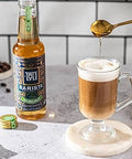 Barista-style coffee being poured into a glass mug with a bottle of Tate & Lyle syrup in the background.