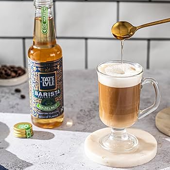 Barista-style coffee being poured into a glass mug with a bottle of Tate & Lyle syrup in the background.